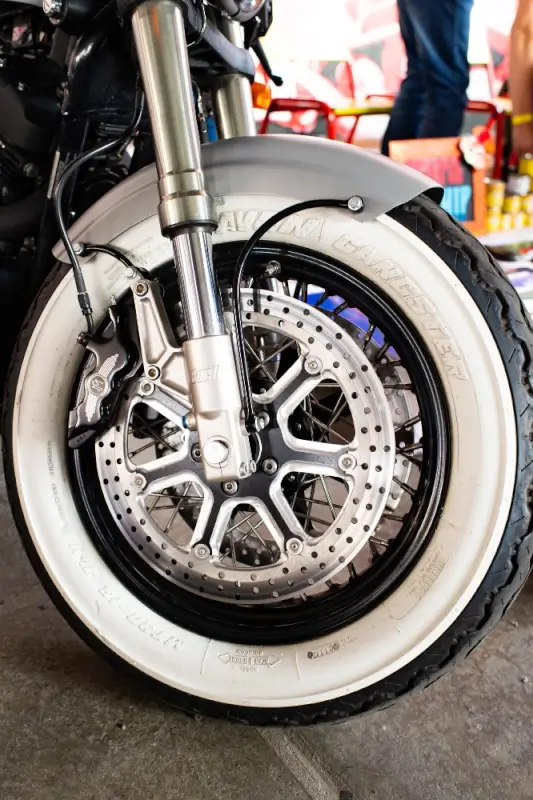 Close-up of a motorcycle’s front wheel showing the brake disc, suspension fork, and whitewall tire, with the tread visible for examining tire wear patterns.