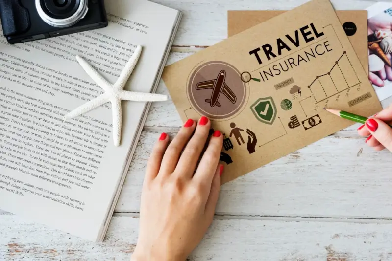 Hand holding travel insurance policy document with airplane icon and coverage symbols next to open guidebook and camera