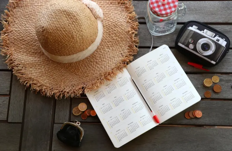 Open travel planner with calendar pages surrounded by straw sun hat, vintage camera, water bottle, coins, and wallet on wooden table