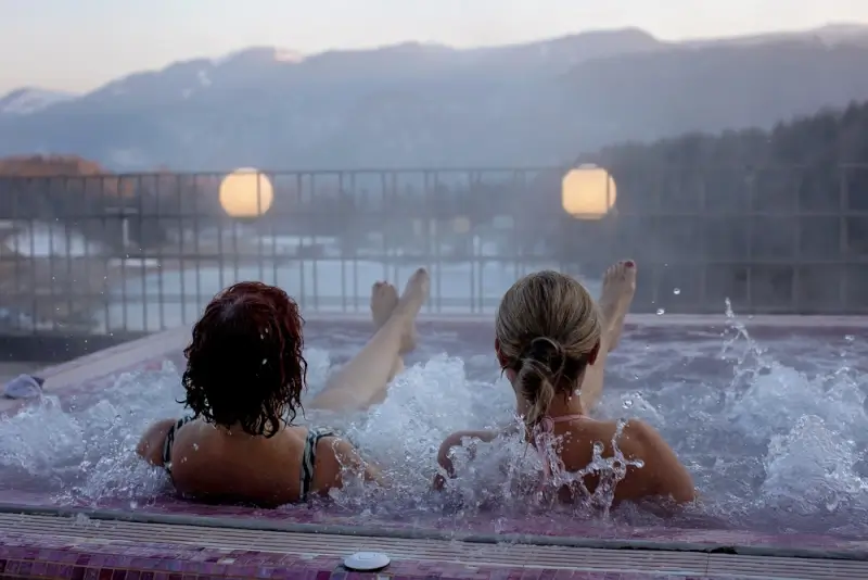Two women relaxing in an outdoor hot tub at dusk, enjoying warm bubbling water with mountain views in the background.