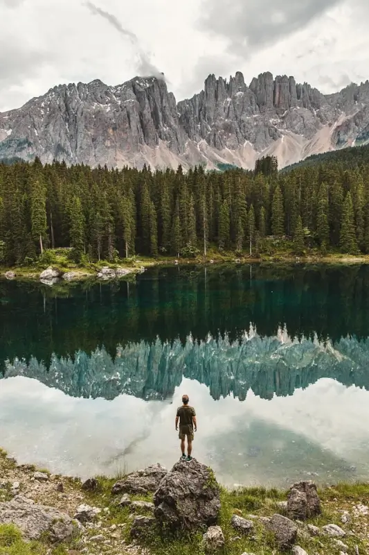 Hiker standing at alpine lake edge with jagged Dolomites peaks reflecting in turquoise water surrounded by pine forest