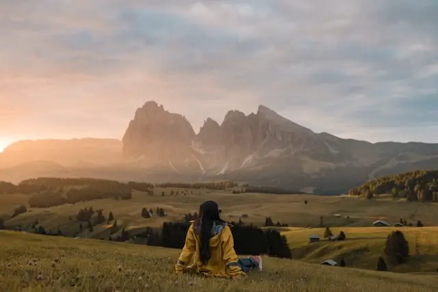 Italian alps hiking through rolling meadows as trekker rests on hillside viewing distant limestone peaks at golden hour