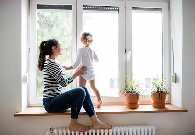 Mother and daughter sitting by sunny window with white radiator beneath windowsill in bright modern home