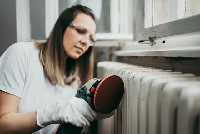 Woman wearing gloves using power sander to refinish white home radiator for year-round heating maintenance