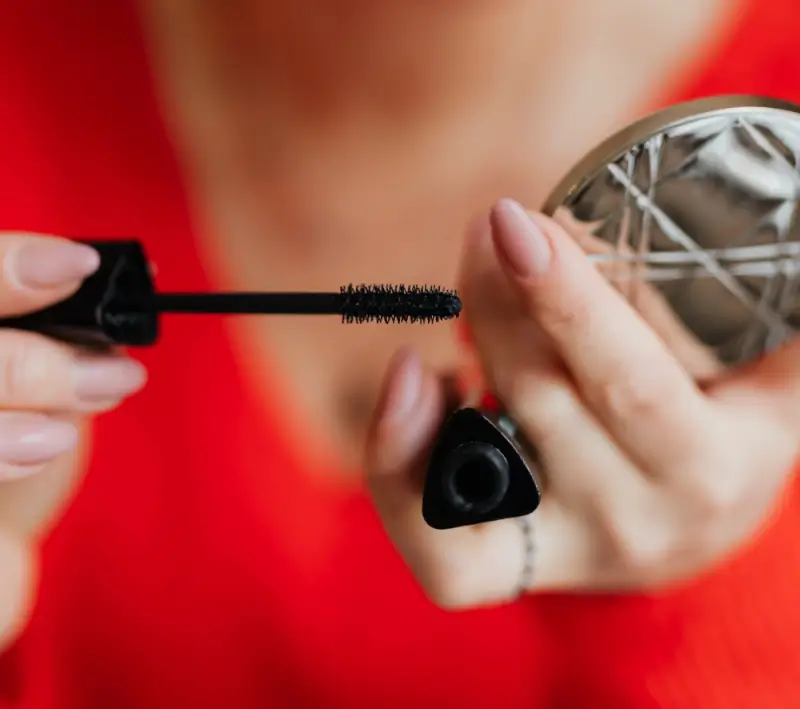 Woman applying mascara while holding compact mirror against red background showing everyday eye makeup routine