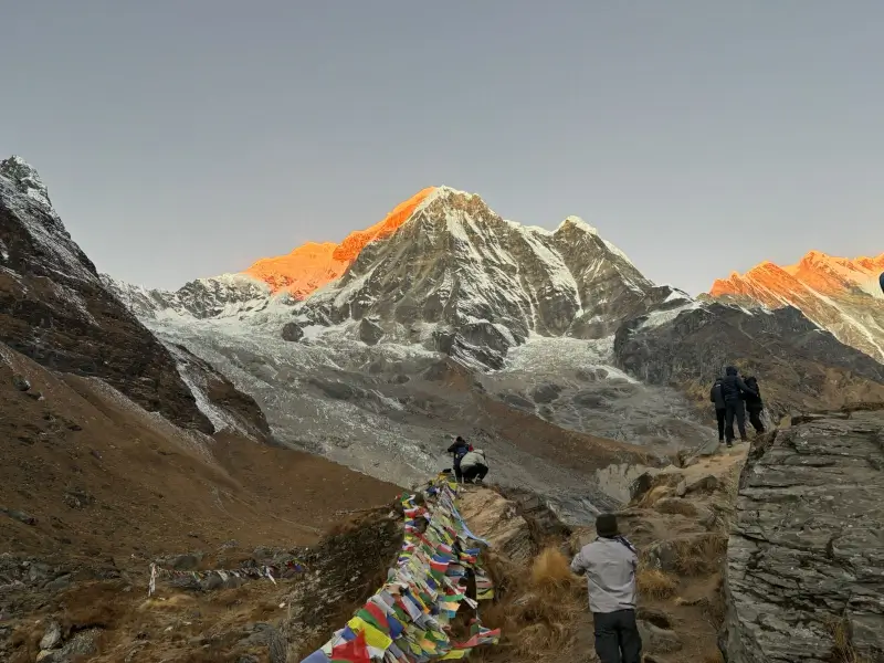 Trekkers walking along the Annapurna Base Camp trail at sunrise, with snow-covered Himalayan peaks and colorful prayer flags in Nepal.