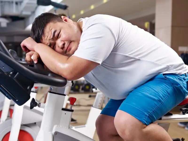 Exhausted man resting on exercise bike showing challenges of weight loss despite lifestyle changes