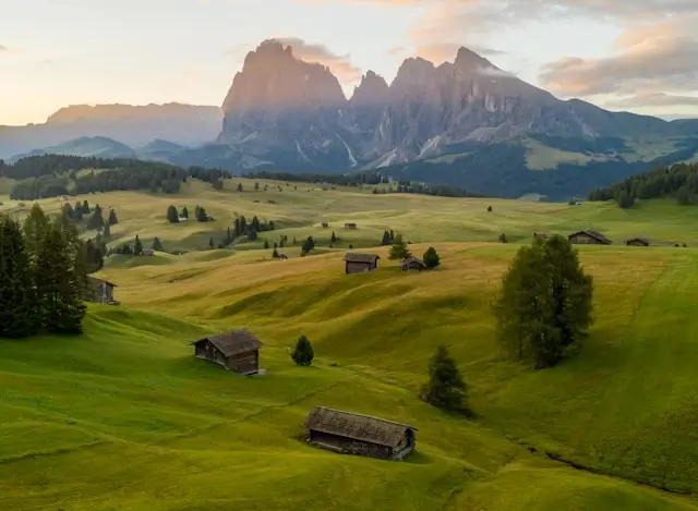 Traditional wooden rifugios scattered across green alpine pastures with dramatic Dolomites mountain backdrop during sunset