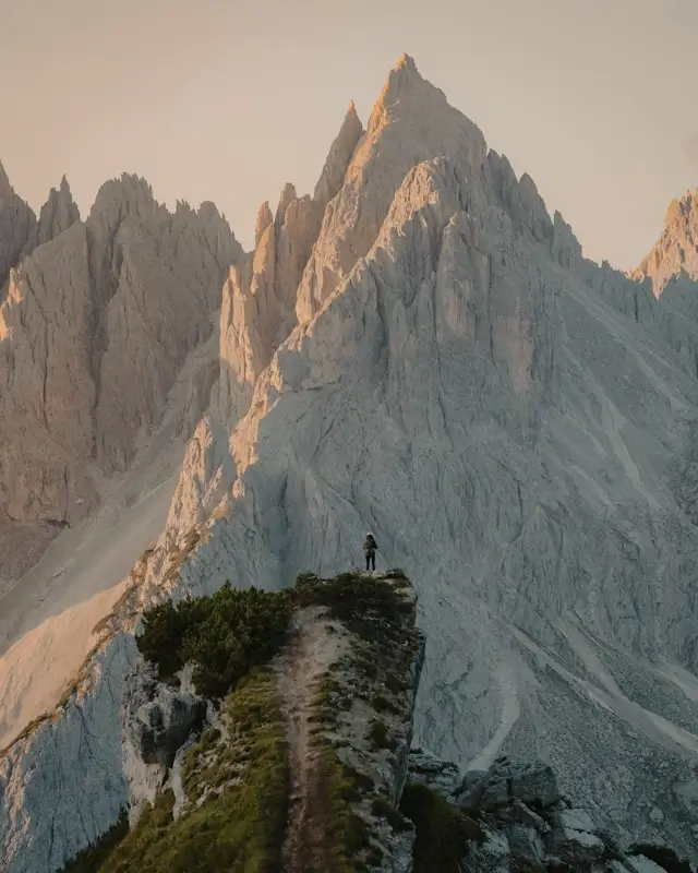 Trekker standing on narrow ridge path with towering pale limestone spires showing enrosadira alpenglow in Italy
