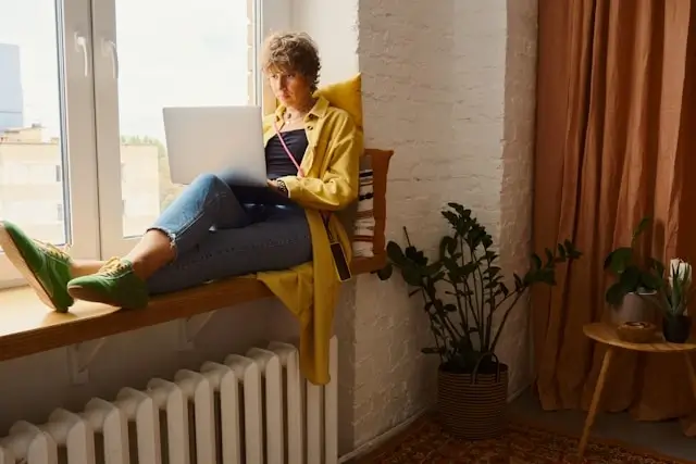 Woman working on laptop by sunny window with white radiator providing comfortable indoor heating throughout the year