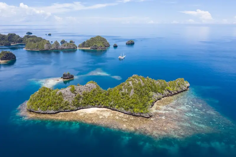 Liveaboard vessel anchored among limestone karst islands in Wayag, northern Raja Ampat, with shallow reefs and deep blue channels visible from above