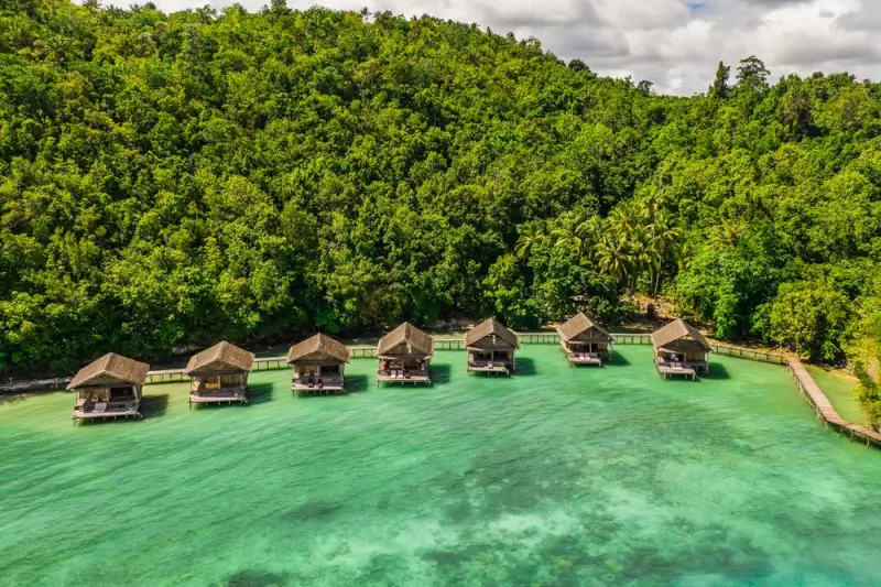 Overwater bungalows at a Raja Ampat resort surrounded by tropical jungle and turquoise lagoon, offering land-based alternative to diving liveaboards