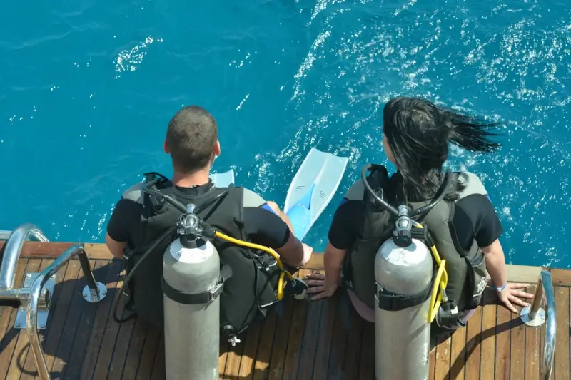 Two scuba divers with tanks and gear preparing to enter turquoise ocean water from dive boat platform
