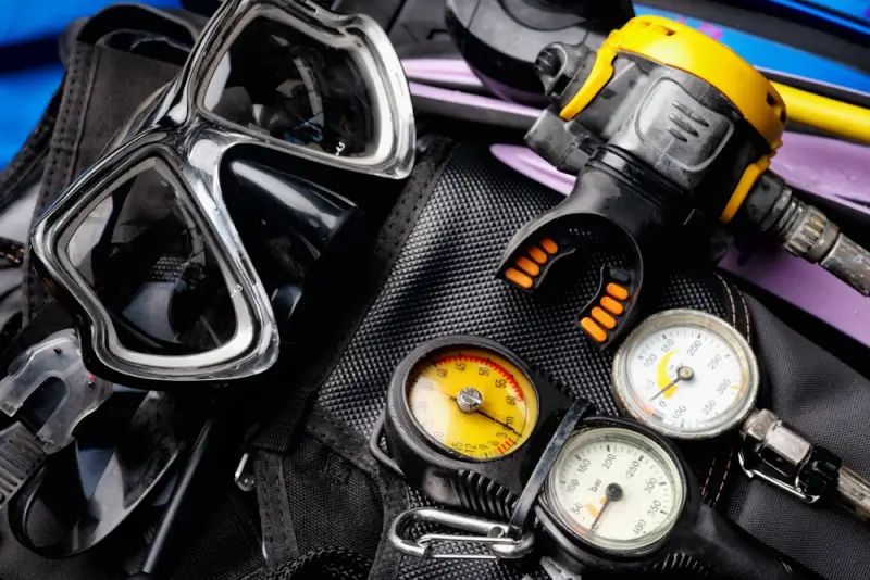 Scuba diving equipment including mask, regulator, pressure gauges, fins, and underwater flashlight laid out on black surface