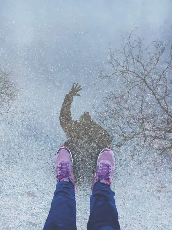 Person standing in pink sneakers looking at reflection in winter puddle with bare trees