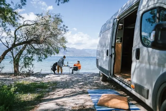 A campervan parked at a lakeside campground in spring with two people relaxing by the water surrounded by mountains