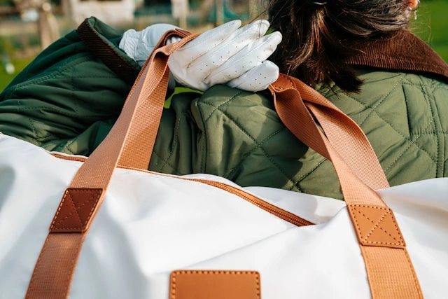 A woman in a green quilted jacket carrying a white canvas duffel bag with brown leather straps over her shoulder outdoors.