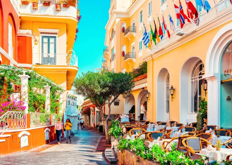 Colorful street in Anacapri with pastel buildings, outdoor cafe seating, and international flags hanging from balconies