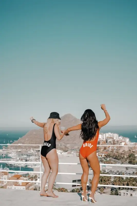Bride and bridesmaid in matching swimsuits celebrating pre-wedding celebrations on rooftop balcony with coastal view