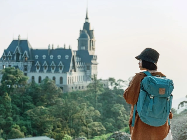 A woman wearing a bucket hat and carrying a teal backpack gazing at a grand castle surrounded by lush greenery.