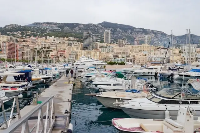 Visitors walking along marina docks in Monaco harbor with luxury yachts moored alongside Mediterranean coastline and hillside buildings