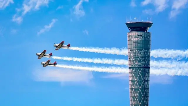 Three aerobatic planes flying in formation past air traffic control tower with white smoke trails