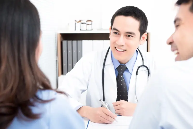 A smiling doctor in a white coat taking notes during a consultation with two patients in a clinic