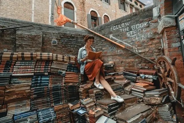 Woman reading a book surrounded by stacks of books on a Venice canal stairway — language learning through travel