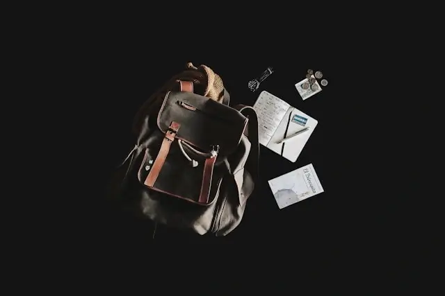 A dark leather and canvas travel backpack with brown straps laid flat against a black background, surrounded by a notebook, coins, a watch, and a small booklet.