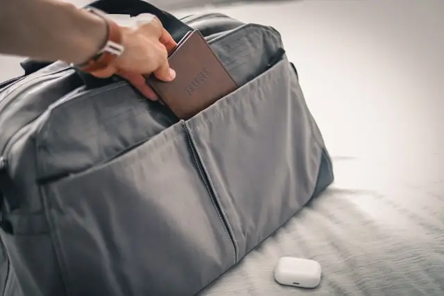 A hand placing a passport into the front pocket of a grey travel duffel bag, with wireless earbuds case resting nearby on a bed.
