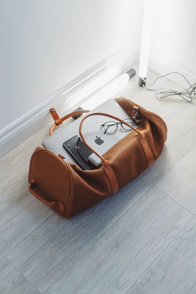 A brown leather travel lifestyle bag line duffel packed with a MacBook, smartphone, and glasses, sitting open on a light wood floor.