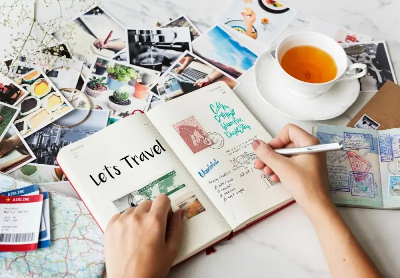 Person writing travel itinerary in journal with photos, passport stamps, tea, and trip planning materials