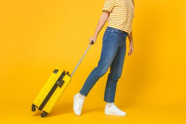 Woman in jeans and white shoes pulling a yellow suitcase against a bright yellow background