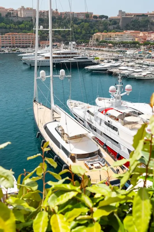 White luxury sailing yacht with tall mast moored in Monaco marina surrounded by superyachts and Mediterranean coastal architecture