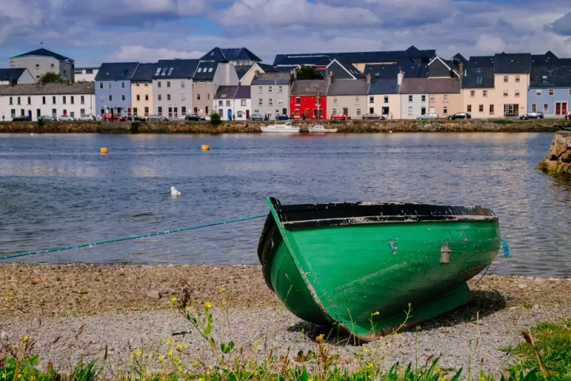 A green wooden boat resting on the rocky shore with Galway's colorful waterfront buildings in the background