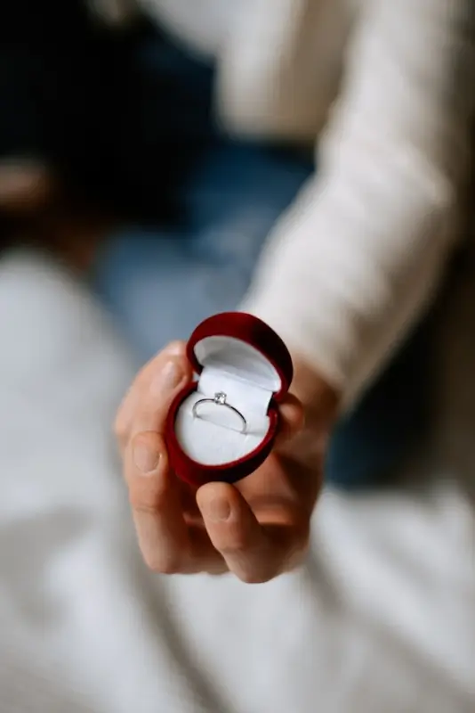 A hand holding a red heart-shaped ring box with a diamond solitaire engagement ring inside