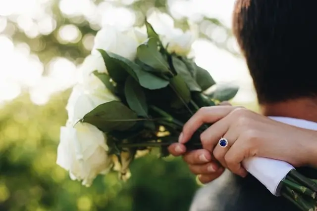 A woman wearing a sapphire halo engagement ring while holding a bouquet of white roses