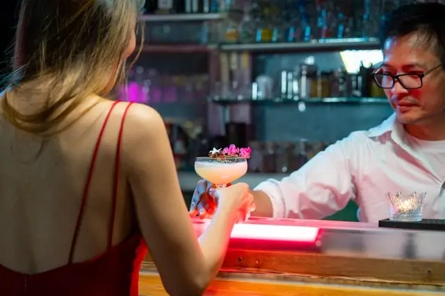 A woman in a red dress receiving a cocktail from a bartender at a dimly lit bar