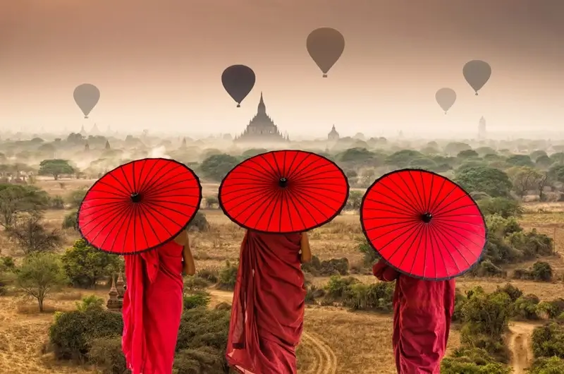 Three Buddhist monks with red umbrellas overlooking ancient temples and hot air balloons over Bagan, Myanmar