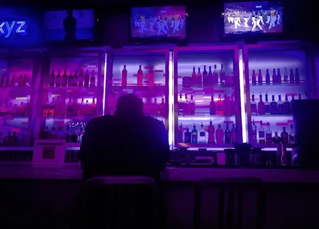 Solo travel at night captured in a single frame — one person seated at a glowing purple bar, surrounded by backlit bottles and screens