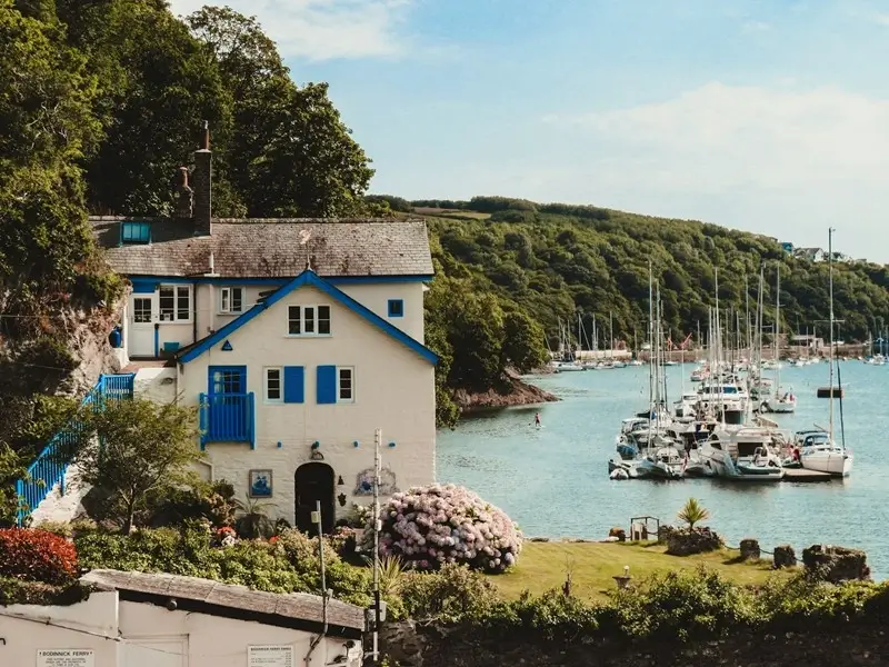 A charming white and blue cottage overlooking a tranquil marina with sailboats and lush green hills in Cornwall