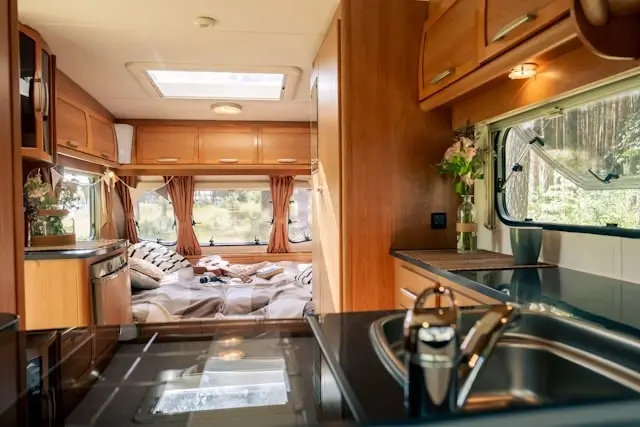 cozy RV interior with a bed, kitchen sink, and wooden cabinets surrounded by nature views