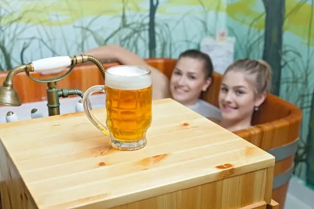 Two women enjoying a beer spa as a fun way to relax in Prague, soaking in a wooden tub with a cold beer mug in the foreground