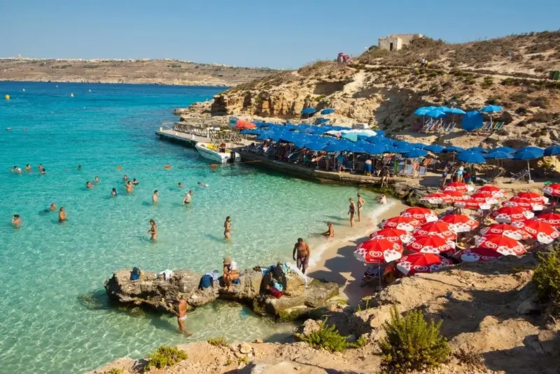 Swimmers and sunbathers at Blue Lagoon Malta with crystal-clear turquoise water, rocky shoreline, and colorful beach umbrellas on a sunny day