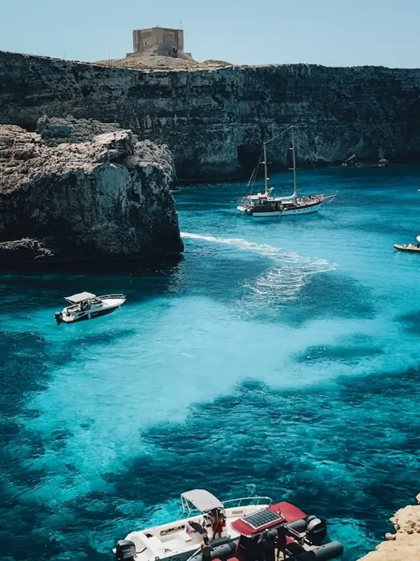 Sailboats and motorboats floating on vivid turquoise water near the rocky cliffs of Comino island with a historic watchtower in the background
