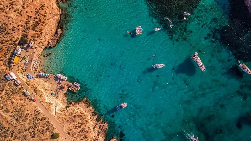 Aerial view of boats anchored in a clear turquoise lagoon surrounded by rocky coastline and a small waterfront area on Comino island