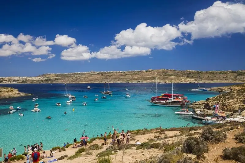 Crowds of visitors and multiple boats gathered along the sandy shore of Comino island on a bright sunny day with vivid turquoise water