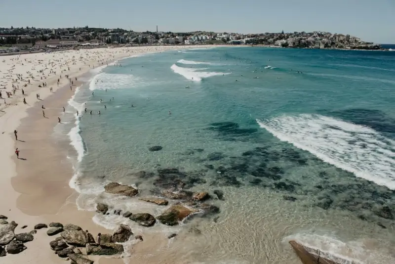 Panoramic view of Bondi Beach in Sydney, Australia, with clear turquoise waters, white sandy shores, and beachgoers scattered along the coastline on a sunny day.