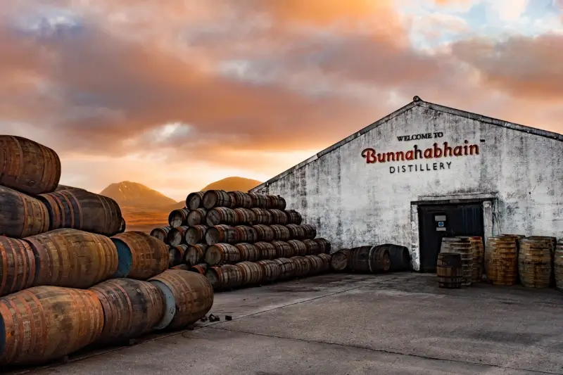 Bunnahabhain Distillery warehouse entrance surrounded by rows of aging oak barrels under a dramatic sunset sky