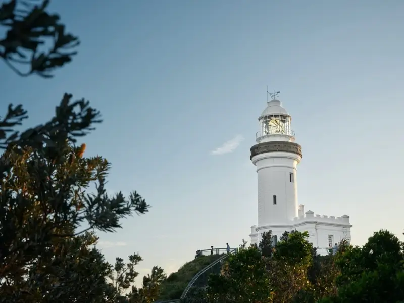 Byron Bay Lighthouse glowing at dusk, framed by native Australian coastal shrubs against a soft pastel sky, with visitors strolling along the path below.